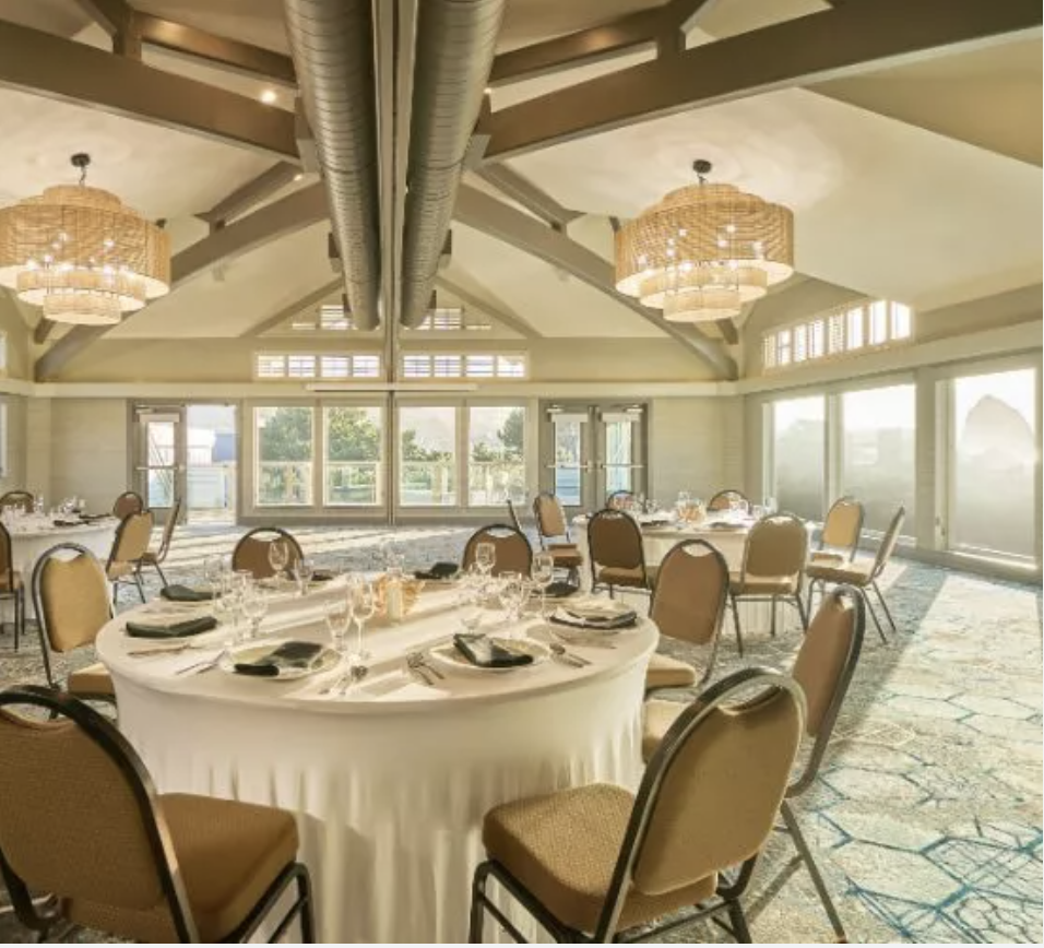 Interior reception space with round tables and floor-to-ceiling windows at Surfsand Resort, softly lit by rattan chandeliers.